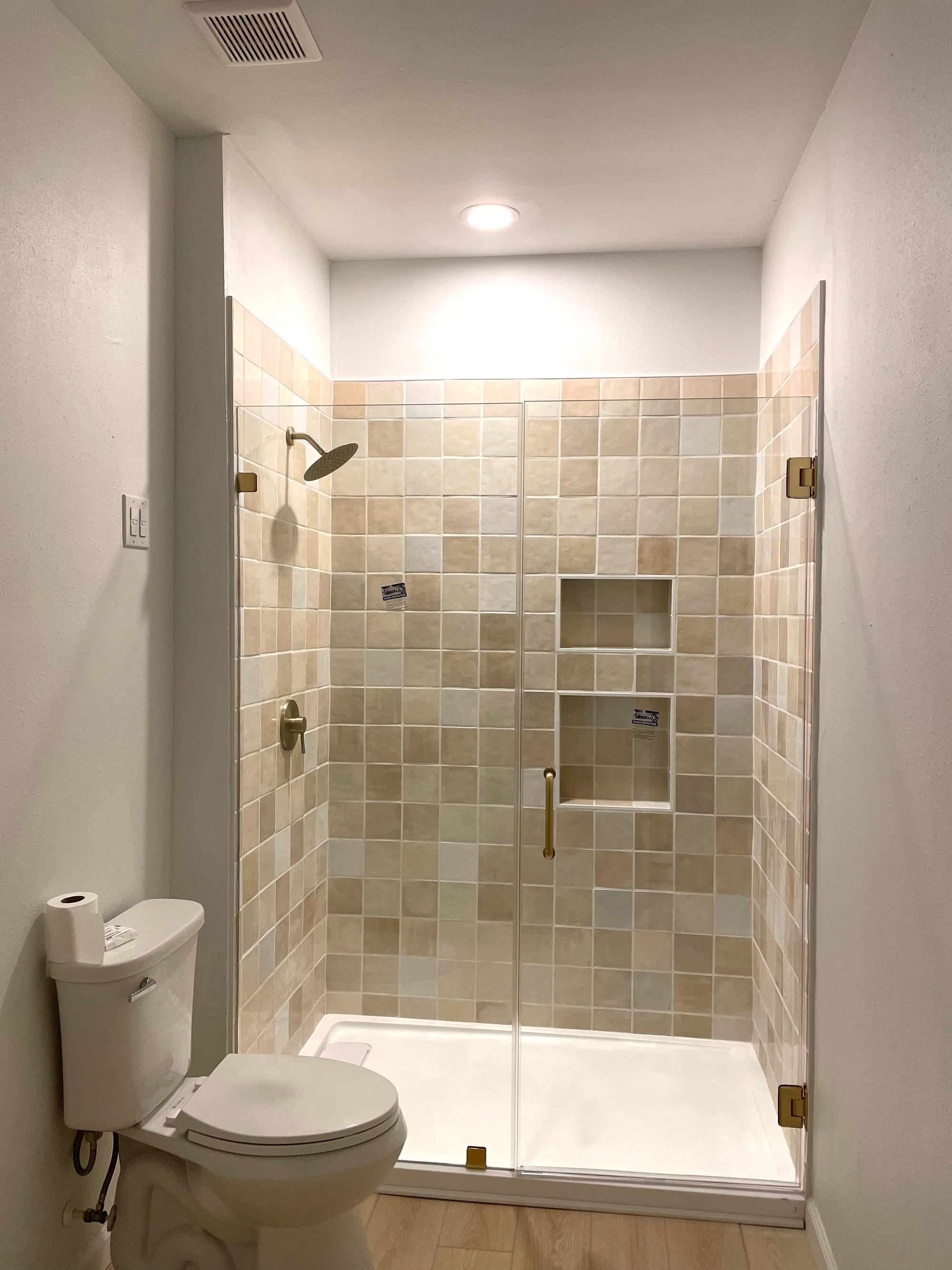 The same alcove as a walk-in shower, tiled floor to ceiling in warm sand-toned square tile with two recessed niches, brass fittings and a frameless glass door.