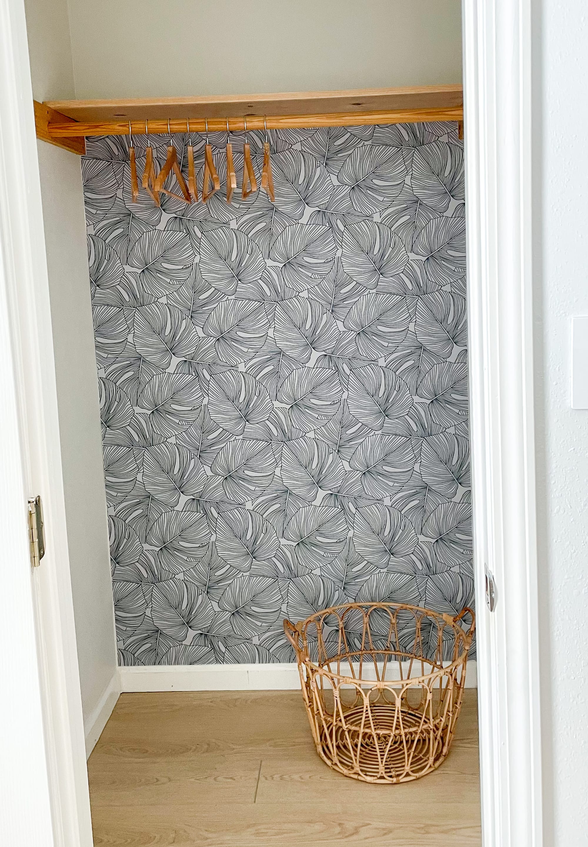 The same closet papered in a large monstera-leaf print, with the timber shelf and rod refinished, oak-look flooring and a rattan basket.