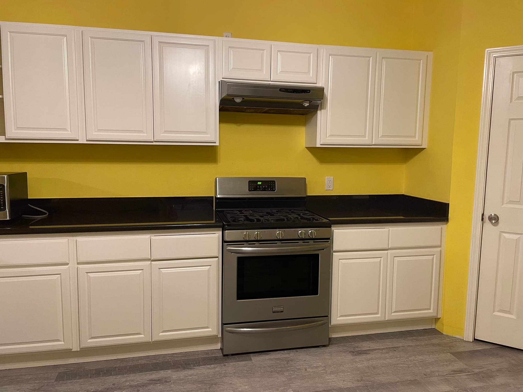 The kitchen with bright yellow walls, white raised-panel cabinets, black counters and a range hood over the cooker.